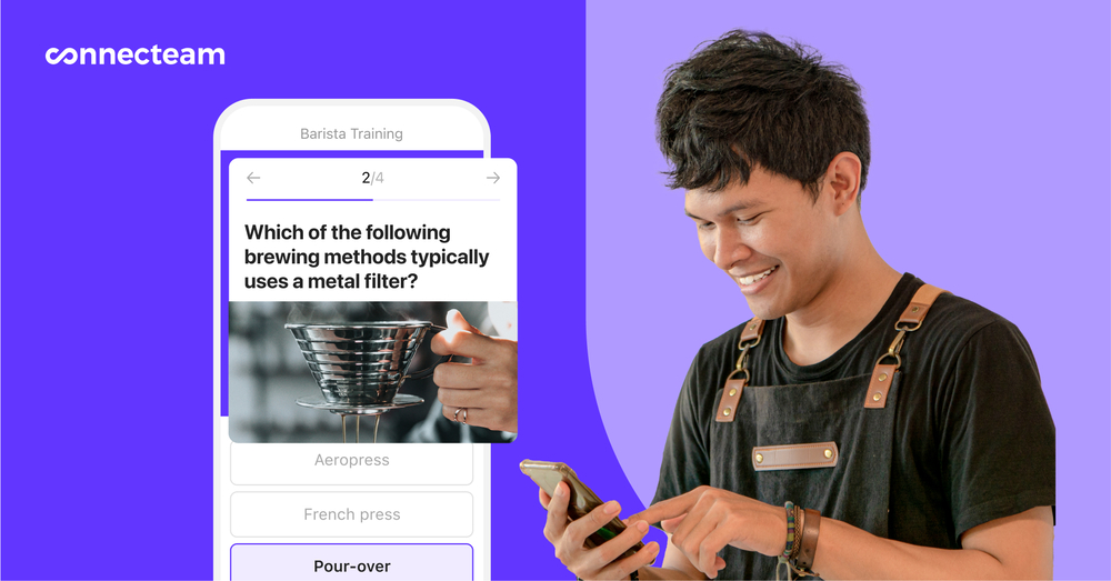 Seorang barista dengan celemek sedang melihat aplikasi Connecteam Training & Onboarding di ponselnya.