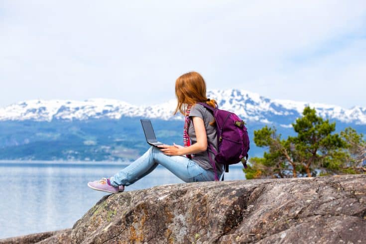 Seorang wanita duduk di atas batu dan menulis blog di komputer laptop; di latar belakang adalah pegunungan yang tertutup salju.