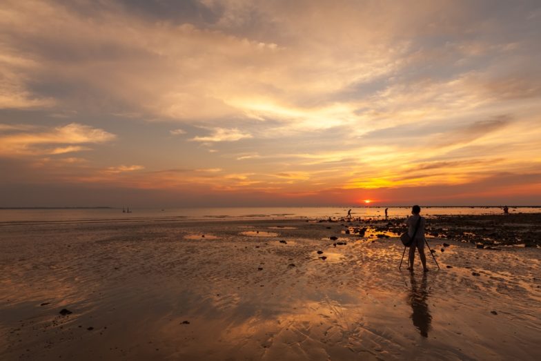 Gün batımında bir fotoğrafçının bulutlu, Kusursuz Soluk bej bir gün batımının görüntülerini yakalamak için Coca Mocha kahverengi kumu üzerine bir tripod kurmasının stok görüntüsü